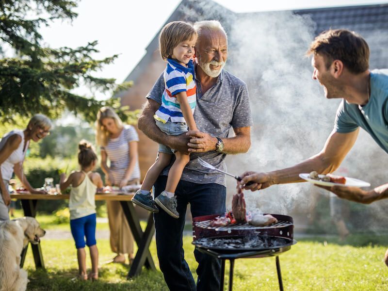 Family gathered around a grill in a sunny backyard, sharing food and a child on an adult’s hip.