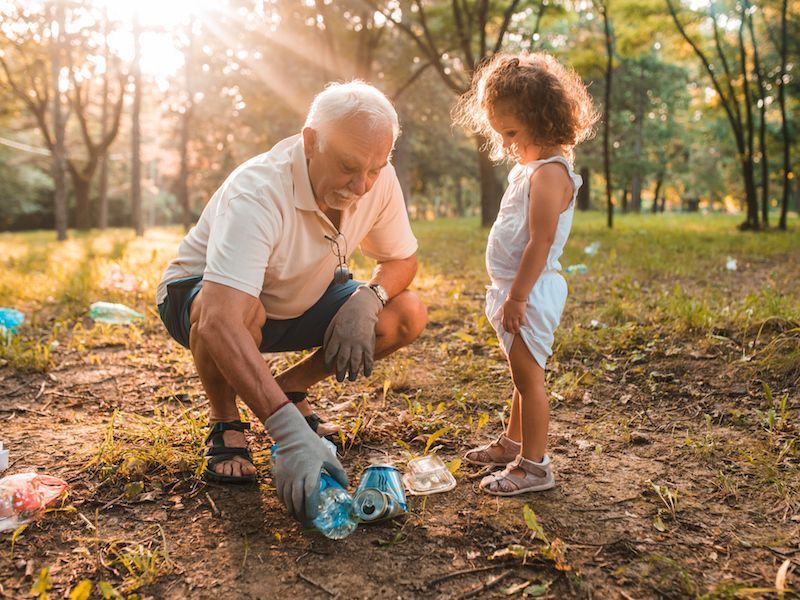 An older man and young child pick up litter in a sunlit park at sunset.