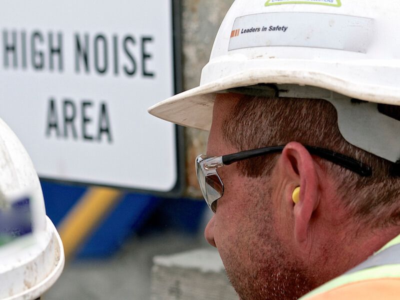Worker in hard hat and safety glasses near a “High Noise Area” sign