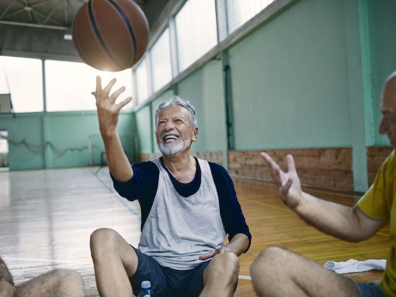 Older man smiling and reaching for a basketball in an indoor gym, with another player nearby