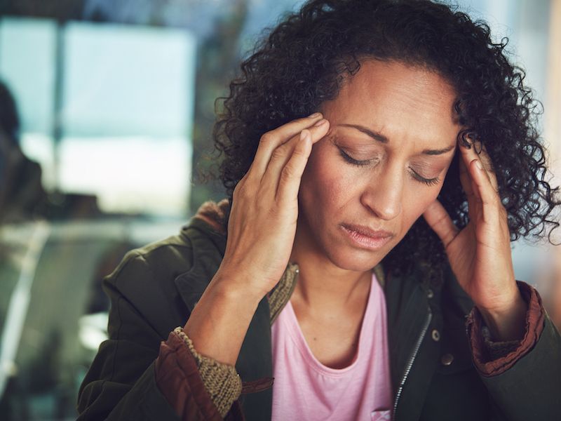 Person in a jacket and pink shirt holds their head with eyes closed, looking stressed indoors.