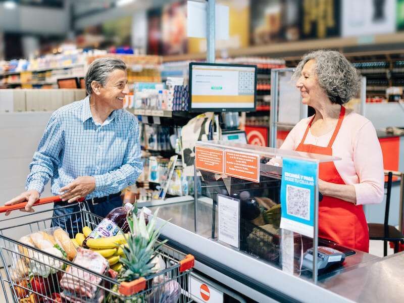 Man and cashier smiling at supermarket checkout with a cart of groceries