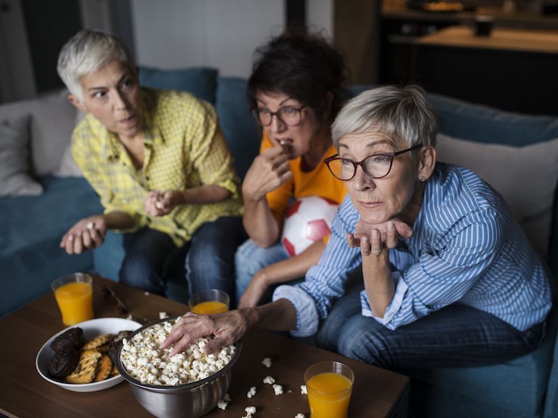 Three friends on a couch eating popcorn and snacks, watching TV together in a living room.