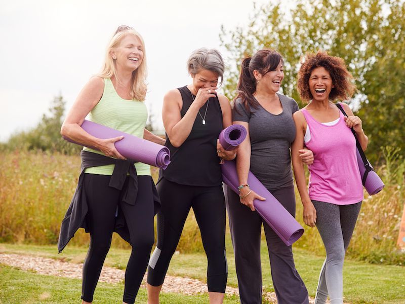 Four women walking outdoors, smiling and holding rolled yoga mats on a path.