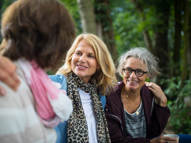 Two smiling people outdoors, talking with a third person in the foreground, in a green park setting.