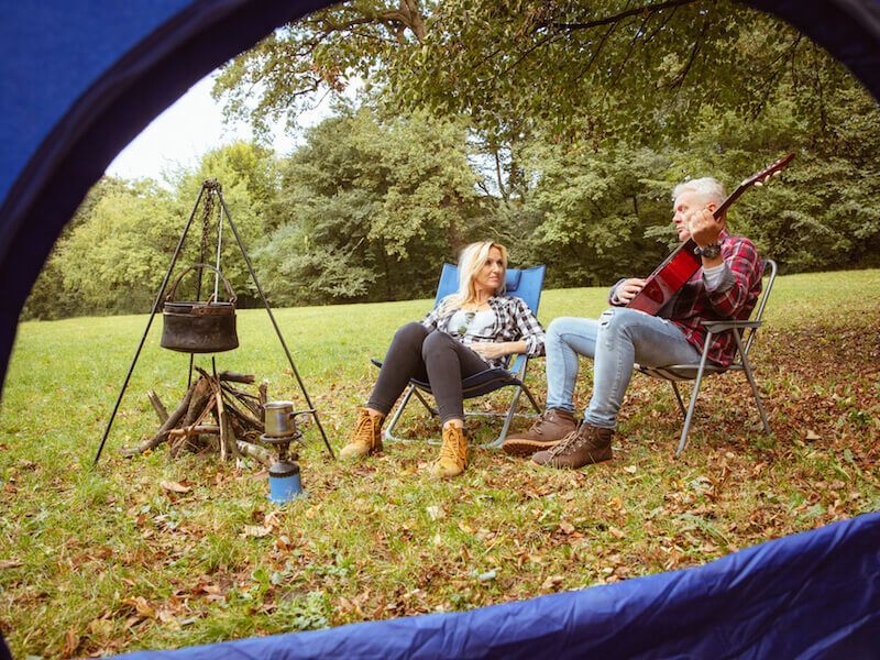 Two people sit in camping chairs under a tent, one playing guitar beside a campfire.