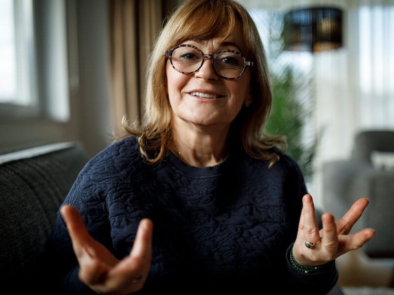 Smiling woman with glasses gesturing while seated on a couch in a bright living room