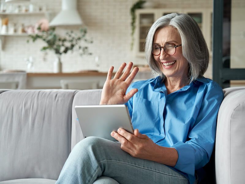 Smiling person in blue shirt waving during a laptop video call on a couch