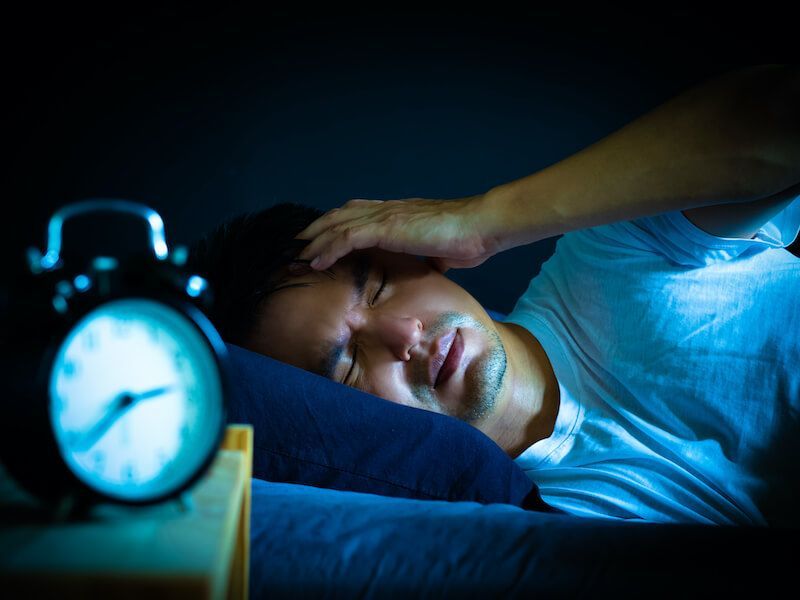 Person lying in bed with a hand on their head, eyes closed, next to a glowing alarm clock in the dark.