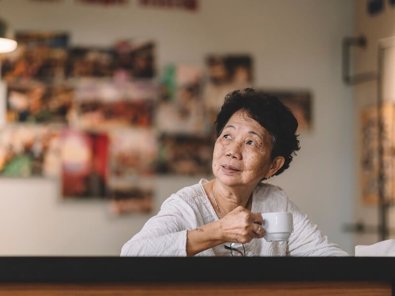Person sitting at a table holding a cup, looking upward in a softly lit room with photos on the wall