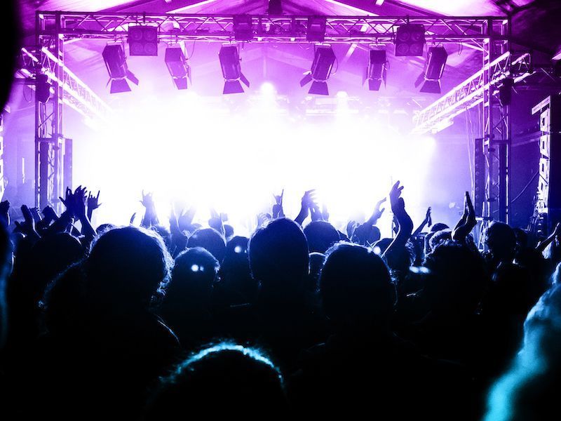Crowd with raised hands at a purple-lit concert stage with bright white lights blasting from the center