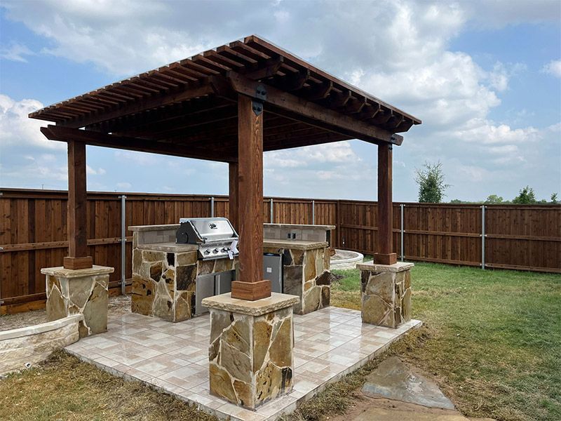 Outdoor stone patio with a wooden pergola, built-in grill, and bar seating in a fenced backyard