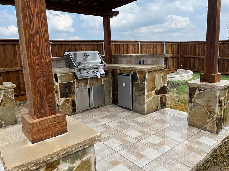 Covered outdoor patio with built-in stone grill and countertop under a wooden pergola, bordered by a fence.