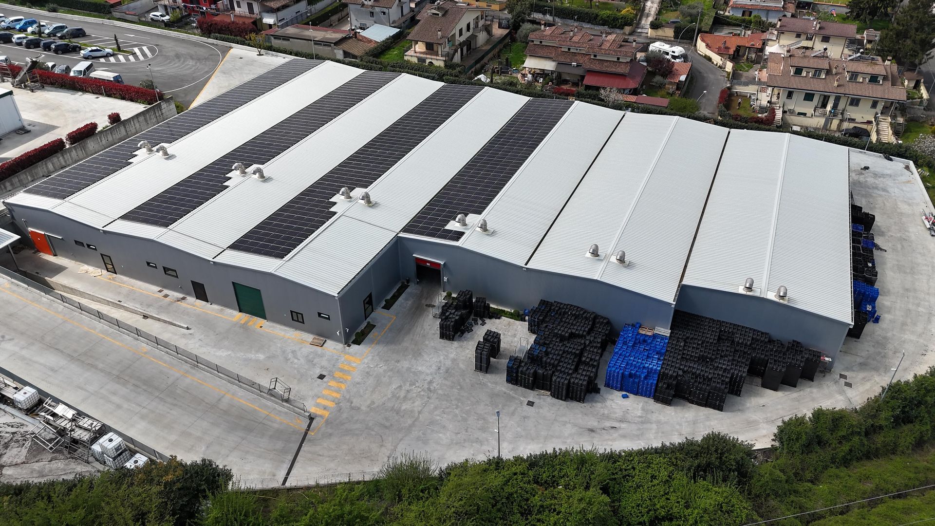 Vista aerea di un edificio industriale con pannelli solari sul tetto. Tetto a strisce grigie e bianche, container scuri all'esterno.