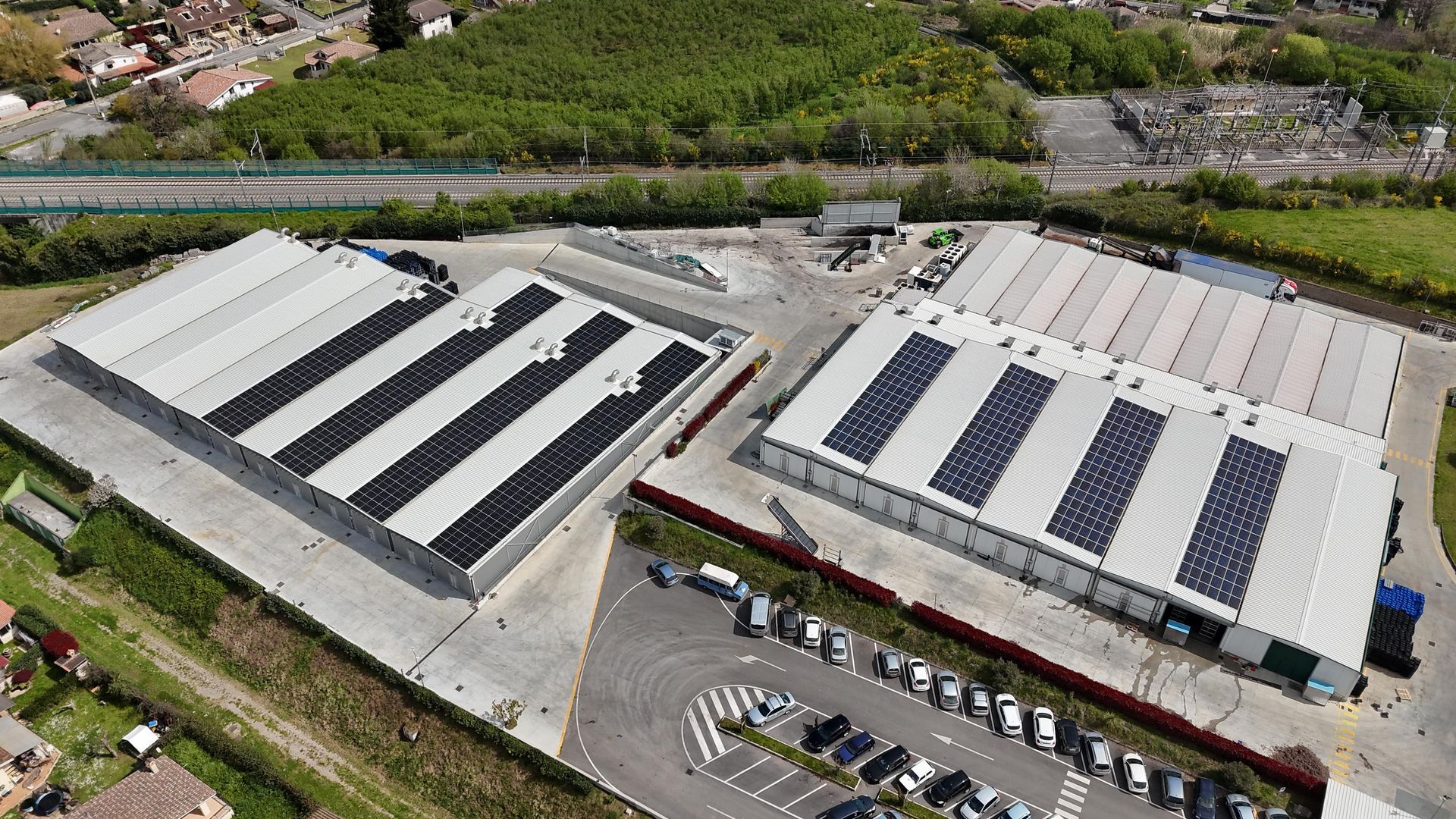 Vista aerea di edifici industriali con pannelli solari sui tetti, parcheggio e vegetazione circostante.