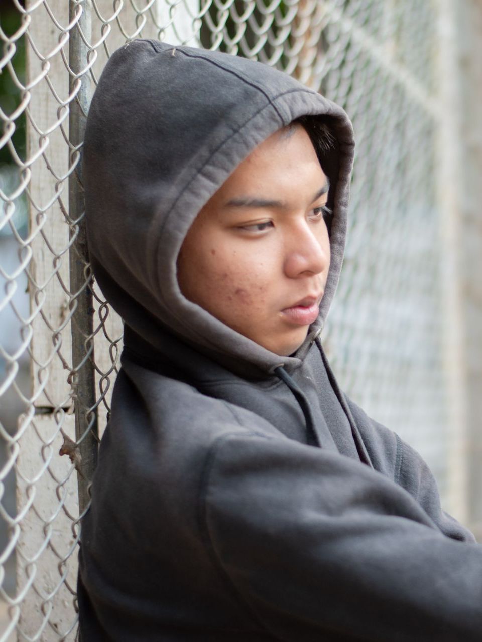 A young man in a hoodie leaning against a chain link fence