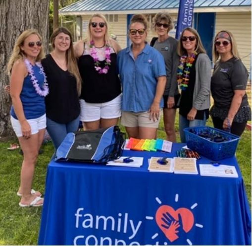 Six people stand behind a Family Connections table at an outdoor event, with colorful materials and T-shirts displayed.