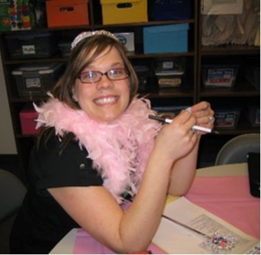 Smiling person wearing a pink feather boa, seated at a desk with colorful storage bins behind.