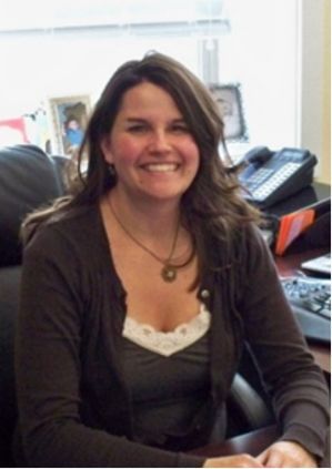 Smiling woman seated at a desk in an office, wearing a dark cardigan and white top.