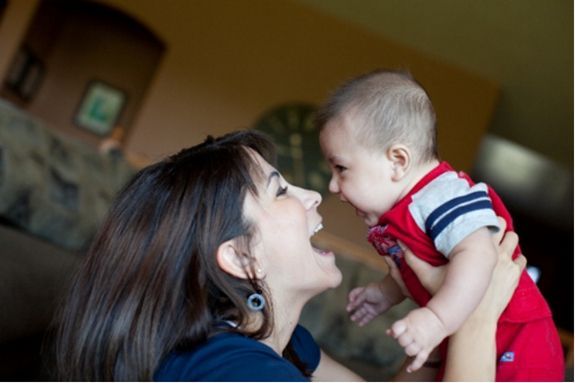 Woman smiling at a baby in red overalls indoors, faces close together