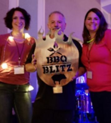 Three people posing indoors with a large “BBQ BLITZ” trophy sign, smiling at an event.