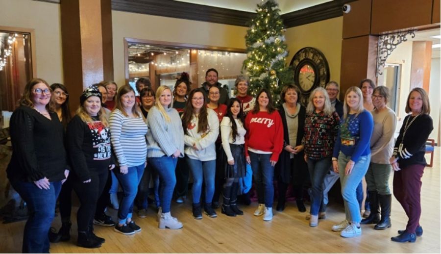 Group of people posing indoors in front of a decorated holiday wreath and tree, smiling at the camera