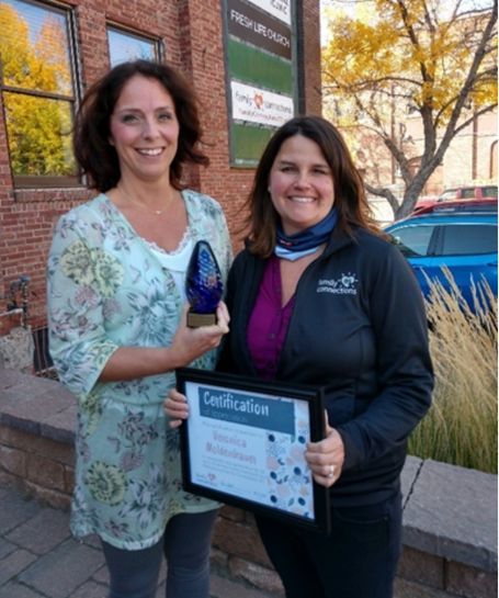 Two women holding an award plaque and glass trophy outdoors near a brick building.
