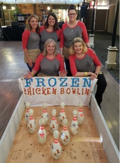 Five women behind a frozen chicken bowling game, with a sign and chicken-shaped pins on the lane.