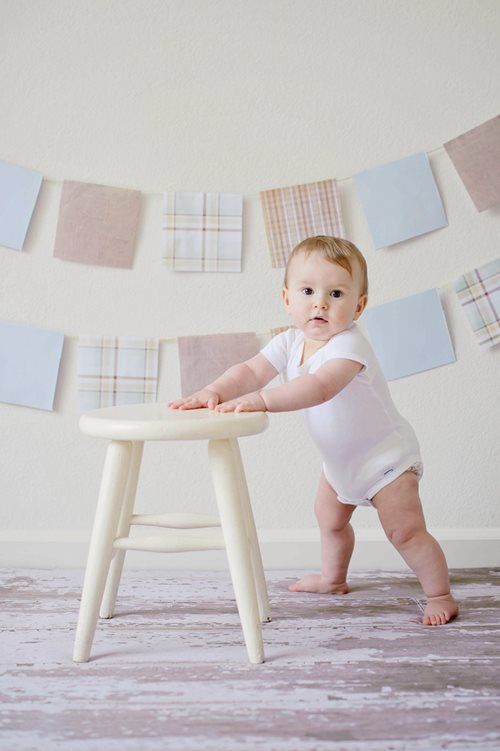 Baby in white onesie standing by a stool in a pastel nursery with hanging fabric banners