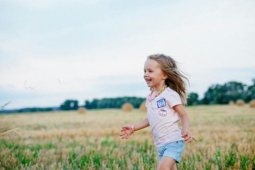 A person with long hair smiles while running through a harvested wheat field under a pale sky.