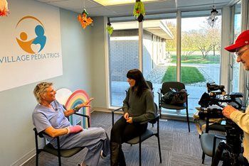 A person in blue scrubs with a pink hand cast talks to another seated person as a videographer films in a pediatrician’s office.