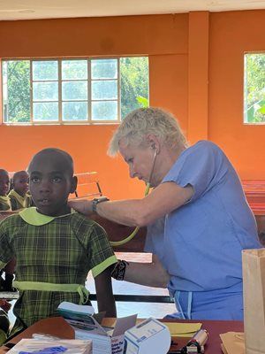 A healthcare provider in blue scrubs uses a stethoscope to examine a child in a green plaid school uniform.