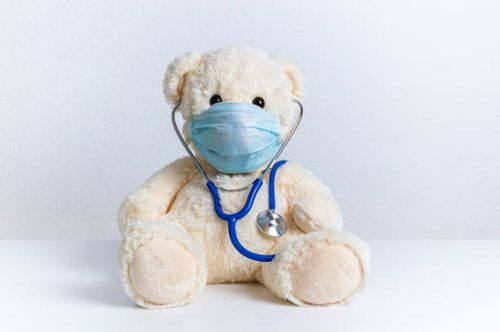 A light-colored teddy bear wearing a blue medical face mask and a blue stethoscope around its neck.