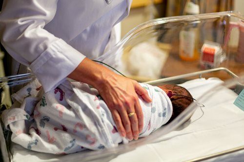 A medical professional in a white coat gently adjusts a newborn swaddled in a patterned blanket inside a hospital bassinet.