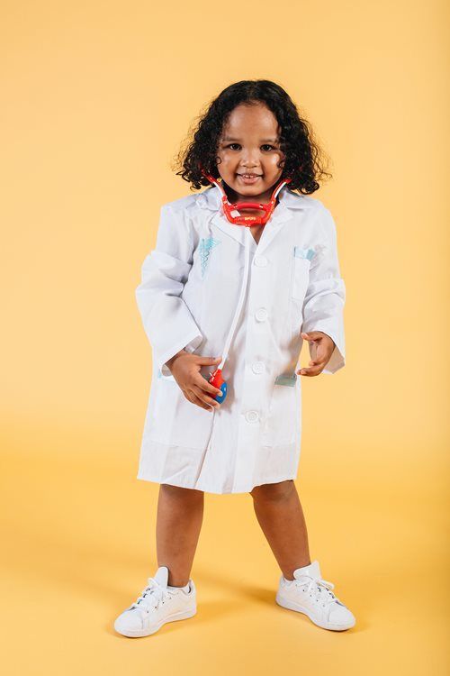 A smiling child in a doctor's costume with a white lab coat and stethoscope standing against a solid yellow background.