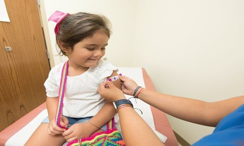 A person places an adhesive bandage on a child's upper arm in a medical exam room.