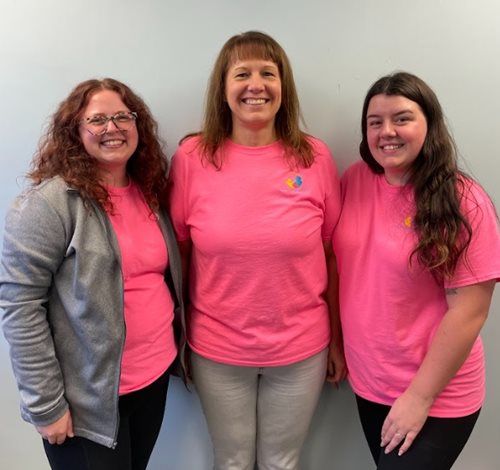 Three people smiling side by side, wearing bright pink t-shirts against a plain light-colored wall.