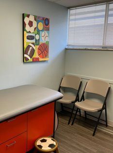 A medical exam room featuring a sports-themed wall art piece, orange storage cabinets, and two folding chairs.