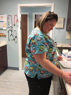 A person in a patterned scrub top writing at a medical office counter, with stethoscopes hanging on the wall behind them.