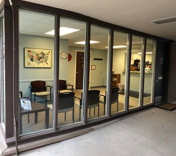 A ground-level view of a brightly lit waiting room through a glass wall, showing chairs, a side table, and a wall map.