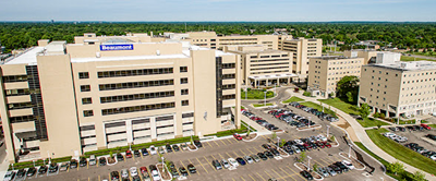 An aerial view of the beige Lakewood Health System hospital complex, featuring multiple wings and an adjacent parking lot.