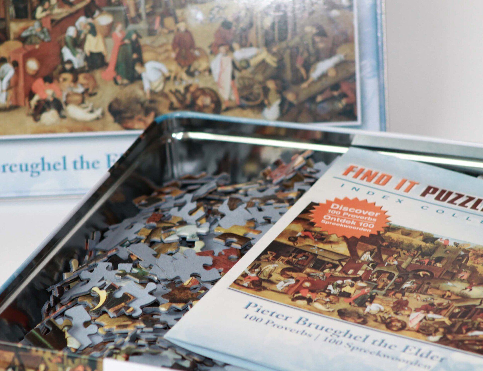 Een metalen puzzelblik gevuld met losse stukjes, met een afbeelding van een schilderij van Pieter Brueghel de Oudere op het deksel en het karton.