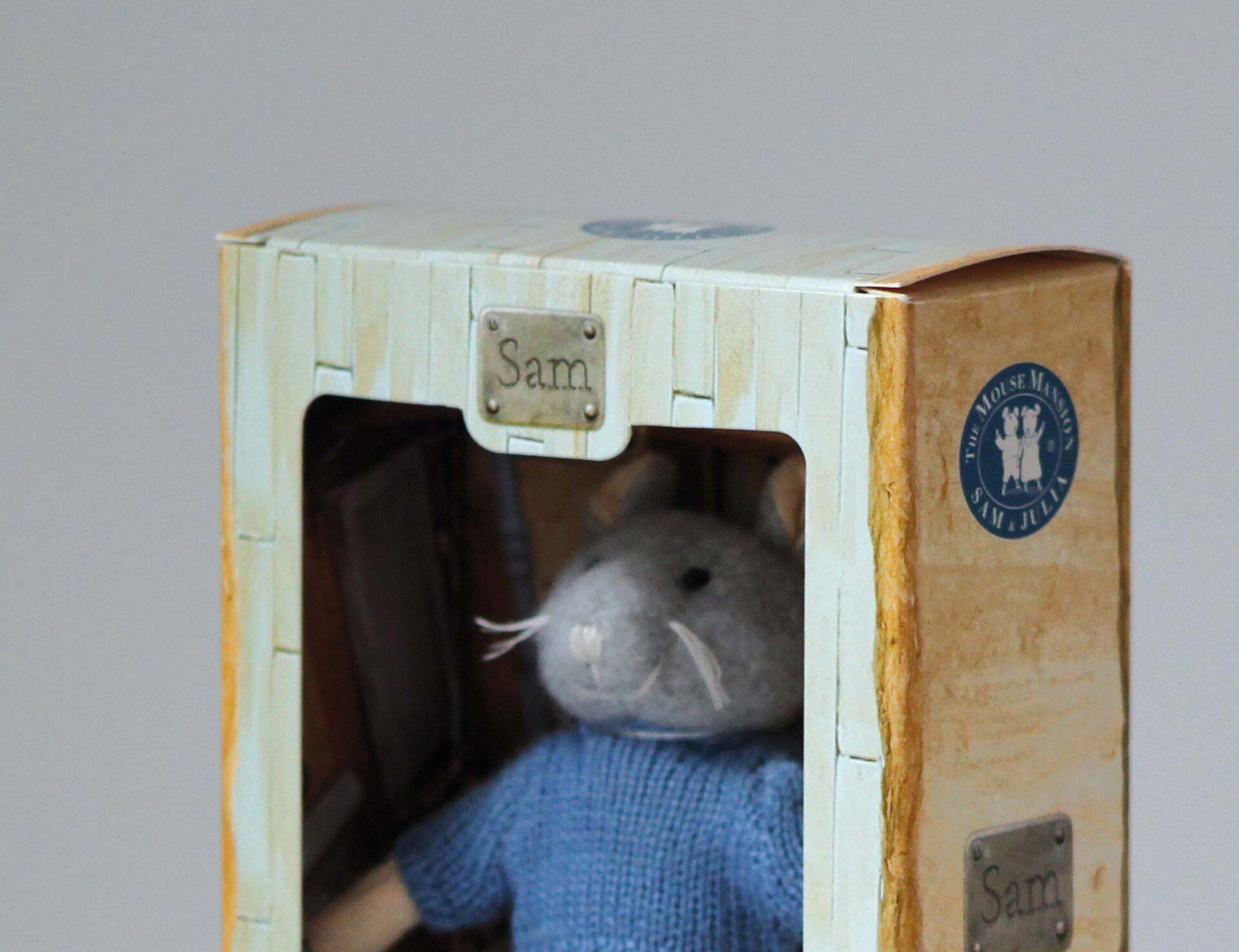 Een grijze vilten muis in een blauwe trui in een lichtgekleurde, rustieke doos met een naamplaatje 