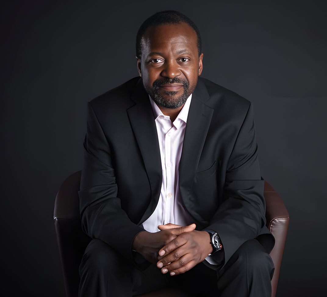 Man in a black suit sits with folded hands, smiling, in front of a dark backdrop.