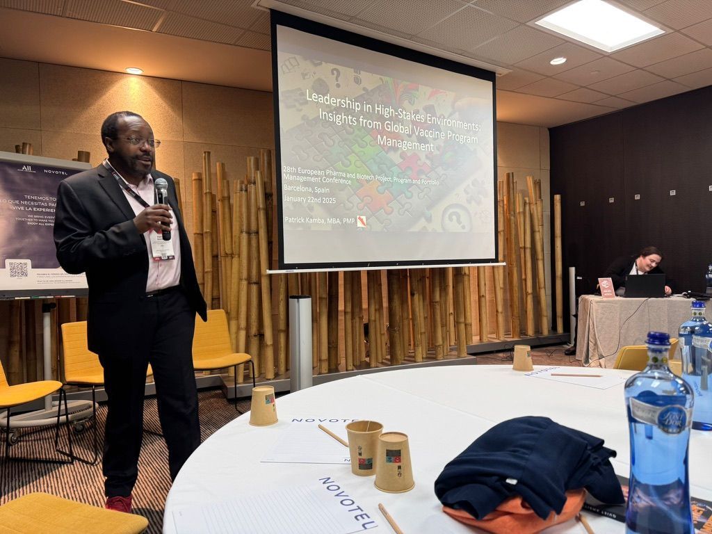 Man presenting at a conference, holding a microphone. Screen with text behind him. Circular table with water bottles and notes.