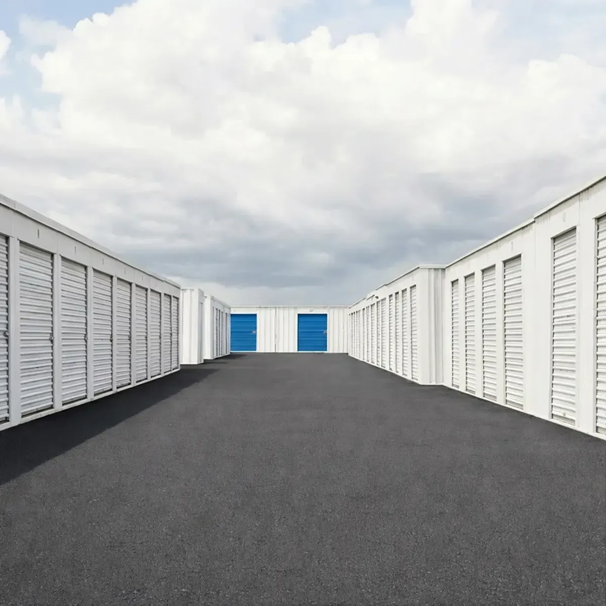 Storage facility aisle between rows of white units, with blue doors in the distance under a cloudy sky.