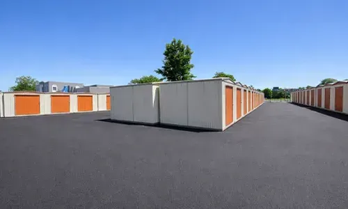 Row of storage units with orange doors in an empty paved lot under a clear blue sky