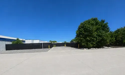 Empty paved lot beside a black fence and trees under a clear blue sky