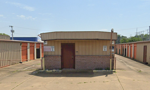 Small brick utility building with tan roof in a concrete courtyard, surrounded by red and beige walls.