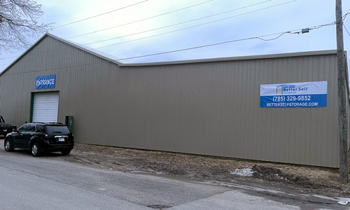 Gray industrial warehouse with a parked black SUV and a blue business sign on the side wall